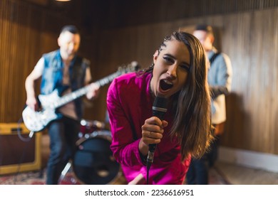 dark-haired enthusiastic woman singing into a microphone and posing for a camera her band playing in the background. High quality photo - Shutterstock ID 2236616951