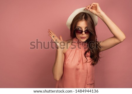 A dark-haired girl in a pink dress, opening her eyes wide, raises a straw hat and points the lodon to the side.