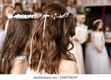 Dark-haired girls with a festive hairstyle and a wreath on their heads are standing in the church. Holy Liturgy. First Communion. Focus delight