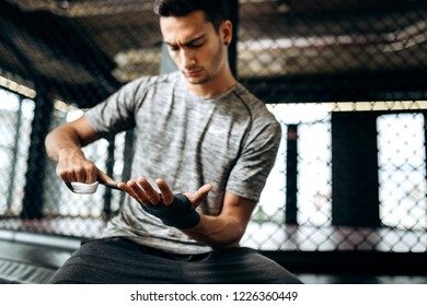 Dark-haired guy dressed in the grey t-shirt wraps a hand bandage on his hand in the boxing gym
