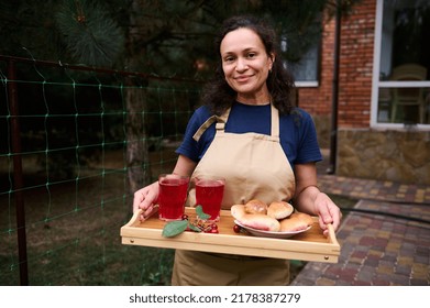 Dark-haired Hispanic Mature Woman In Kitchen Apron Smiles Looking At Camera, Standing In The Home Backyard Garden With A Tray Of Homemade Delicious Cherry Pies And Refreshing Compote In Drinking Glass