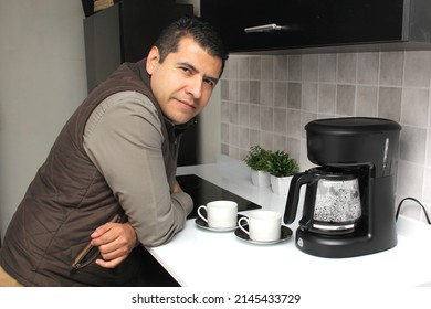 Dark-haired Latino adult man prepares a cup of coffee in a coffee maker to smell and taste at breakfast in the kitchen is thoughtful, worried, thoughtful, depressed
