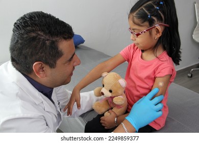 Dark-haired Latino doctor and little girl have a medical consultation in the pediatric office to vaccinate their arm against Covid, chickenpox, diphtheria, influenza, hepatitis, measles, mumps
 - Shutterstock ID 2249859377