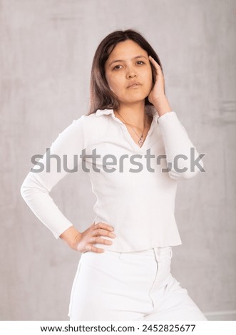 Dark-haired model in light clothes put one hand on waist and coquettishly holds unfastened hair with handbreadth.Studio shot, gray background