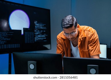 A dark-haired network defence analyst studies an alert bent over the desk in a blue-lit cyber operations centre. The image conveys tracking suspicious activity across enterprise systems.