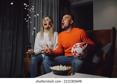 Dark-haired Screaming Enthusiastic Man And Blonde Woman Staring Screen Sports Soccer Victory Goal Watching TV Scattered Popcorn On Comfort Brown Couch. Indoor Dark Side View