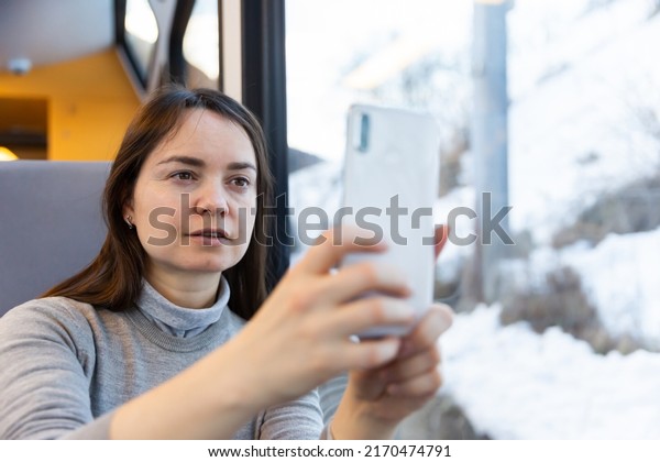 Dark-haired woman enjoying journey in modern\
comfortable train, looking with interest at picturesque scenery\
outside window and taking pictures on\
cellphone