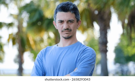 A dark-haired young man looking at the camera in a tree-filled park, with his hands crossed over his chest and a unhappy frown