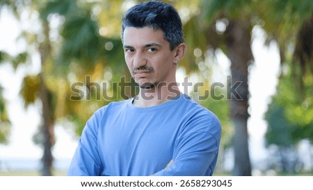 A dark-haired young man looking at the camera in a tree-filled park, with his hands crossed over his chest and a unhappy frown