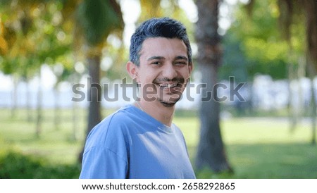 A dark-haired young man smiling at the camera in a tree-filled park