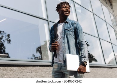 a dark-skinned African American with a laptop in his hands walks down the street against a stele building, in the city center - Powered by Shutterstock