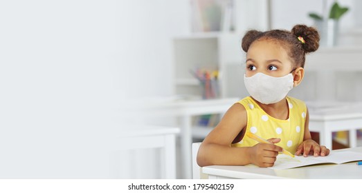 A dark-skinned girl in a medical mask for respiratory protection in a yellow dress listens attentively to the teacher. white background. Banner format - Shutterstock ID 1795402159