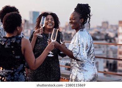 Dark-skinned girl pours champagne into glasses of her multinational friends. Cute happy people having fun at rooftop party. African women in elegant dresses and young man drink wine and laugh. - Powered by Shutterstock