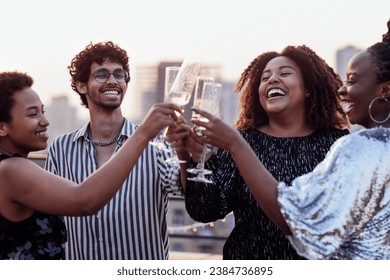 Dark-skinned girl pours champagne into glasses of her multinational friends. Cute happy people having fun at rooftop party. African women in elegant dresses and young man drink wine and laugh. - Powered by Shutterstock