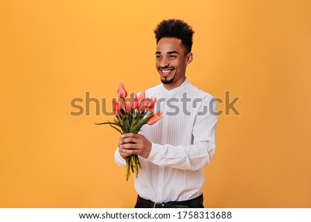 Dark-skinned guy laughs and holds bouquet of flowers on orange background. Charming man in white shirt waiting for date and posing with tulips on isolated backdrop