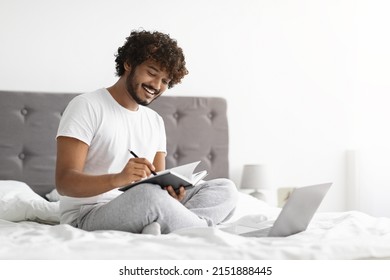 Dark-skinned Guy In Pajamas Sitting On Bed At Home, Using Modern Laptop, Taking Notes And Smiling, Unemployed Young Man Looking For Job Opportunities Online, Side View, Copy Space