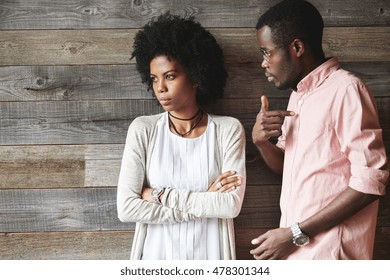 Dark-skinned man standing next to his offended wife with Afro haircut and crossed arms, man looking guilty, pointing finger at him, asking what he did wrong, trying to understand what happened to her