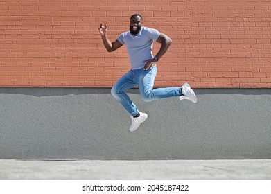 A Dark-skinned Man In A White Tshirt And Jeans Jumping Near The Brick Wall