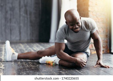 A dark-skinned smiling fitness trainer in a gray T-shirt sits on the floor and does exercises to the side