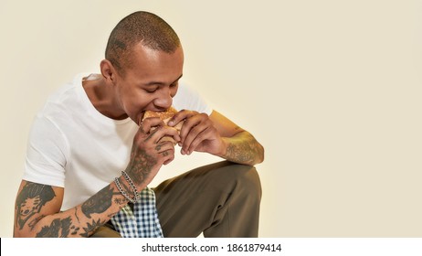 A Dark-skinned Tattooed Man Eating A Bugrer Sitting On His Legs Wearing A White T-shirt On A Light Yellow Background