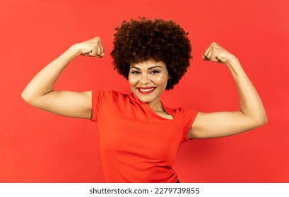 A dark-skinned woman with afro hair poses on a red background. The girl shows the two biceps with expression of encouragement, winner or motivation. Concept of empowered woman, overcoming. - Shutterstock ID 2279739855