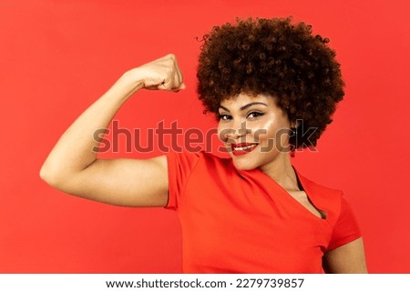 A dark-skinned woman with afro hair poses on a red background. The girl shows a biceps with expression of encouragement, winner or motivation. Concept of empowered woman, overcoming.