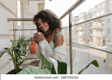 Dark-skinned woman in light-green silk dress drinks coffee on balcony. Attractive lady in white cardigan enjoys espresso on terrace. - Powered by Shutterstock