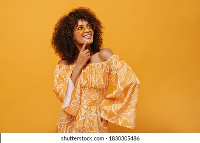 Dark-skinned Woman In Sunglasses Smiling On Isolated Backdrop. Pensive Girl In Summer Clothes Posing On Yellow Background..