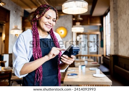 A dark-skinned woman is in uniform working inside a coffee shop. The African girl with colored braided hair is handling a POS terminal. Concept of women working in hospitality. African waitress girls.
