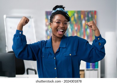 Dark-skinned woman wearing blue clothes glasses stands against office backdrop smiles happy, victory, winning, raises hands up tenses hand muscles gestures strength power successful girl businesswoman - Powered by Shutterstock