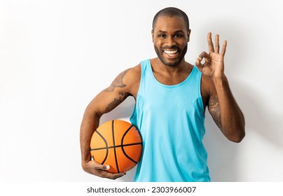 A dark-skinned young man is posing on a white background wearing a tank top and holding a basketball. The man has a cheerful expression,makes the OK gesture with his hand,happy with the match. - Powered by Shutterstock