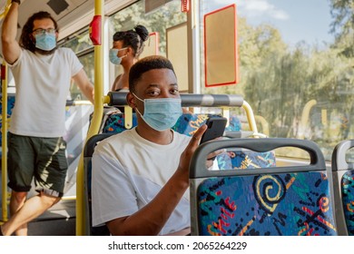 Dark-skinned Young Man Sits On Public Transport Bus Face Mask Phone In Hand, Texting With Friends, Two People In Background Stand Holding Handles Talking To Each Other, Afternoon Returning From Work