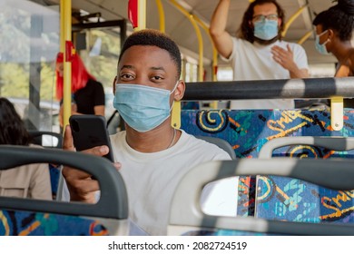 Dark-skinned Young Man Sits On Public Transport Bus Face Mask Phone In Hand, Texting With Friends, Two People In Background Stand Holding Handles Talking To Each Other, Afternoon Returning From Work