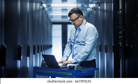 In Data Center IT Engineer Works On Crash Cart Laptop. We See Rows Of Server Racks And One Cabinet Is Open.