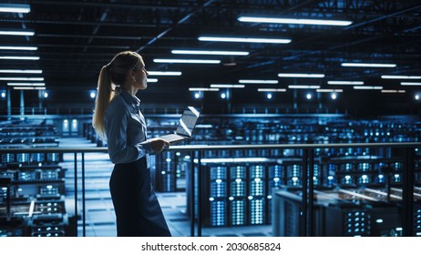 Data Center Female IT Specialist Uses Laptop Computer. Information Technology Engineer Overseeing And Walking Through Cloud Computing Server Farm Maintenance And Monitoring.