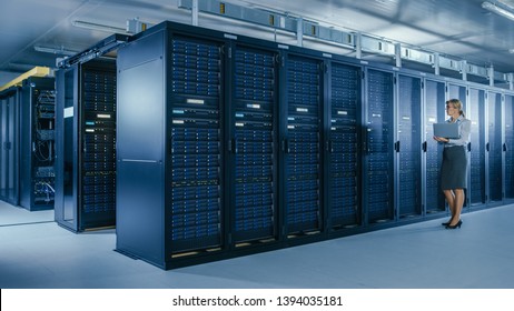 In Data Center: Female IT Specialist Walks along the Row of Operational Server Racks, Uses Laptop to Run Maintenance Programme. Modern High-Tech Telecommunications Operational Data Center.