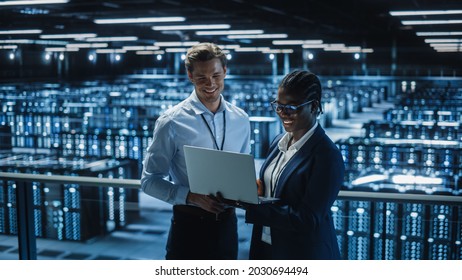 Data Center Female System Administrator And Male IT Specialist Talk, Use Laptop. Information Technology Engineers Work On Cyber Security Protection In Cloud Computing Server Farm.