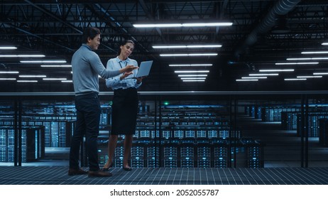 Data Center Female System Administrator And Male IT Specialist Talk, Use Laptop Computer. Information Technology Engineers Work On Cyber Security Network Protection In Cloud Computing Server Farm.