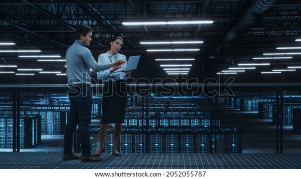 Data Center Female System Administrator and Male IT\
Specialist Talk, Use Laptop Computer. Information Technology\
Engineers work on Cyber Security Network Protection in Cloud\
Computing Server Farm.
