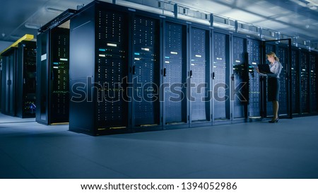 In Data Center: Female IT Technician Stands before Open Server Rack Cabinet, Uses Laptop Computer to Run Maintenance Diagnostics so that Mainframe Works at Optimal Functioning Level.