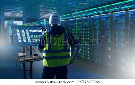 Data center, information technology, engineering. Technician in safety vest and helmet monitors server racks using a control panel, ensuring network security and system performance.