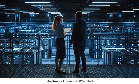 Data Center Male It Specialist And Female E-Commerce Manager Talk, Use Laptop. Big Server Farm Cloud Computing Facility With Information Technology Professionals Working.