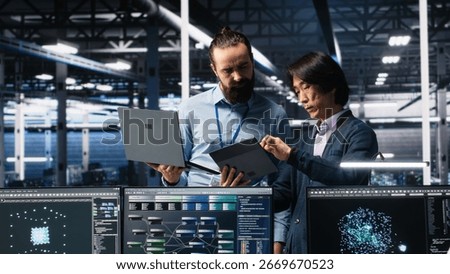 Data center manager supervising developer using device to monitor neural network AI LLMs. Team leader checking progress done by worker overseeing servers powering machine learning processes, camera A