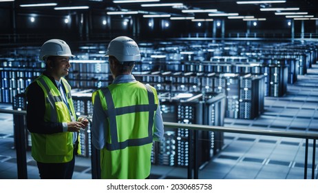 Data Center IT Specialist and System administrator Talk, Use Tablet Computer, Wearing Safety Wests. Server Clod Farm with Two Information Technology Engineers checking Cyber Security.