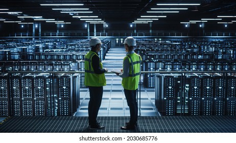 Data Center IT Specialist and System administrator Talk, Use Tablet Computer, Wearing Safety West. Server Cloud Farm Facility with Two Information Technology Engineers checking Cyber Security.