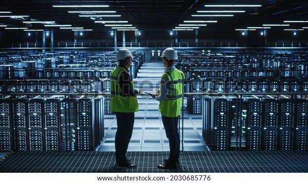 Data Center IT Specialist and System\
administrator Talk, Use Tablet Computer, Wearing Safety West.\
Server Cloud Farm Facility with Two Information Technology\
Engineers checking Cyber\
Security.