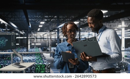 Data center system administrators using laptop to review AI LLM visualizations. African american IT specialists use device to analyze neural networks outputs, ensuring stable gear operations