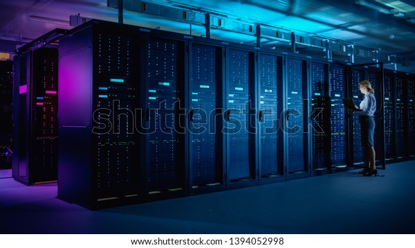 In Data Center: IT Technician Running Maintenance\
Programme on Laptop, Controls Operational Server Rack Optimal\
Functioning. Modern High-Tech Telecommunications Operational Data\
Center in Neon Lights.