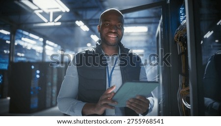 Data center worker uses a tablet to monitor server rack performance, checks network cables, and inspects illuminated hardware in facility filled with servers. Portrait, looking at camera