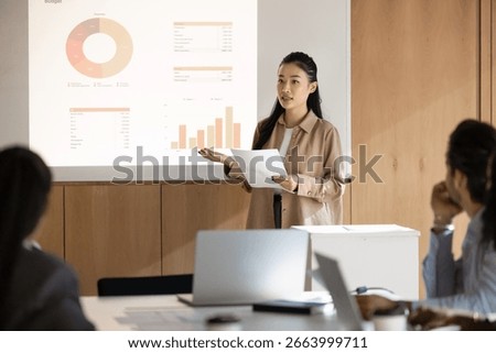 Data driven seminar. Young Asian woman speaker lead interactive presentation in modern meeting room use digital screen to visualize project information in charts graphs give report to diverse audience